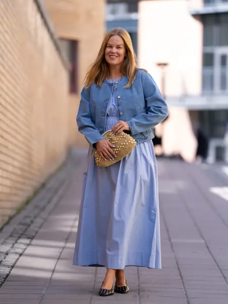 Dame med blå lang kjole, olajakke, sorte flate sko og beige veske med perler.