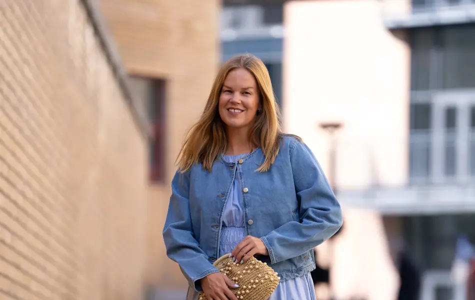 Dame med blå lang kjole, olajakke, sorte flate sko og beige veske med perler.