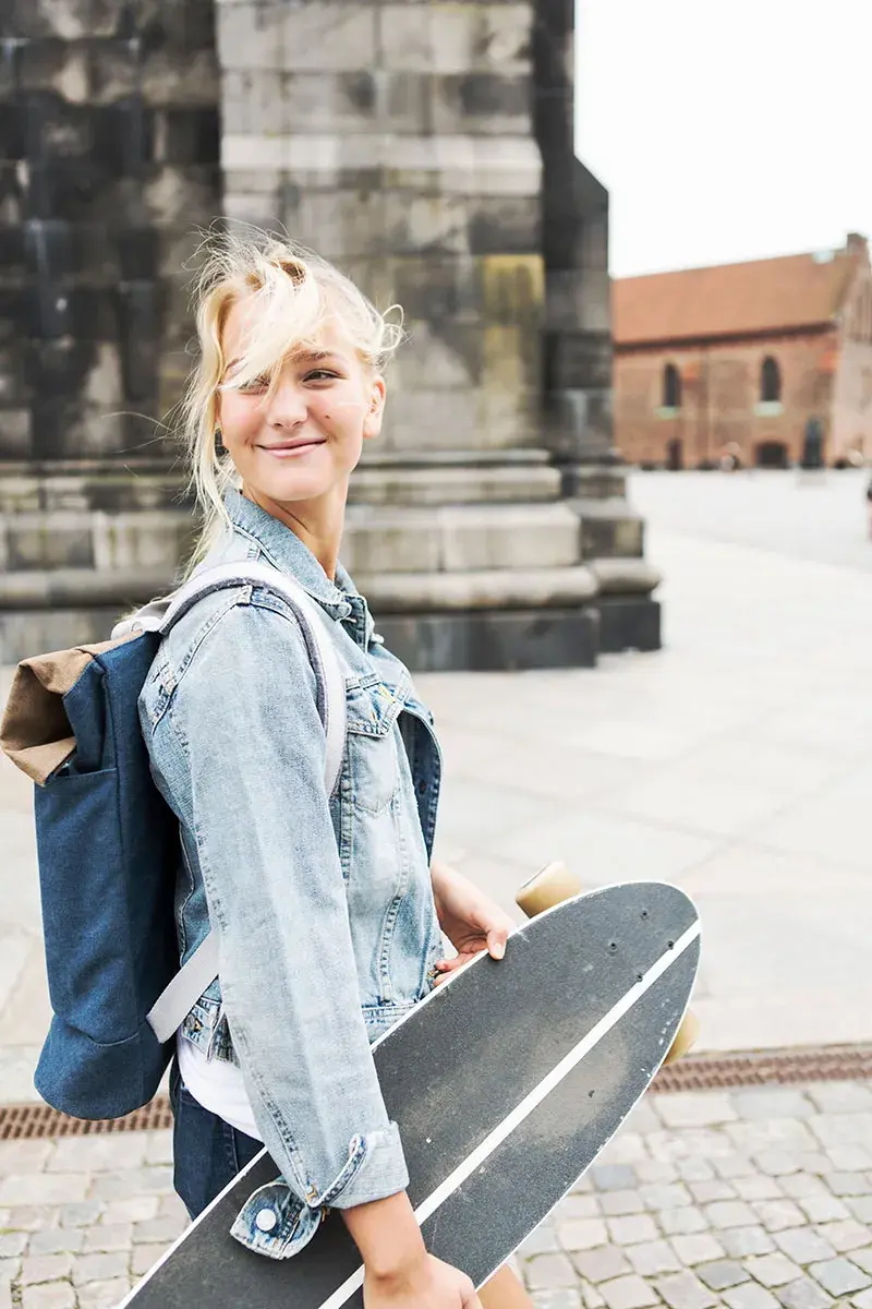 Smilende blond jente som går med sekk på ryggen og skateboard under armen
