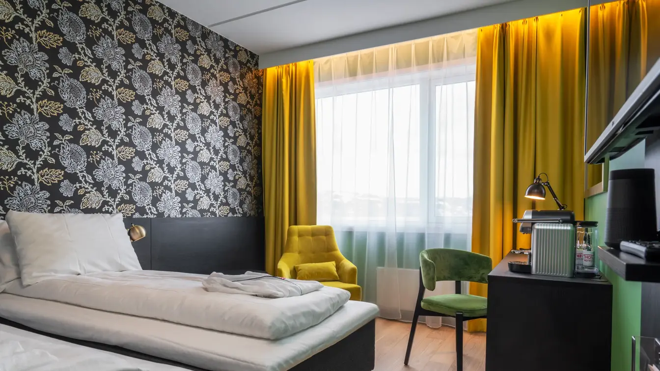 Separate senger, mørk mønstrete tapet med blomster, gule gardiner, gul stol, skrivebord med kaffemaskin og grønn stol i superior twin rom på Thon Hotel Kirkenes
