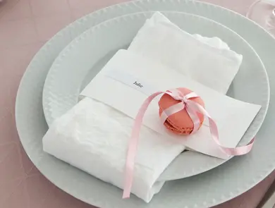 To hvite tallerker, kniv og gaffel, rosa duk og hvit serviett med rosa makron knyttet med silkebånd og en navnelapp hvor de står Julie