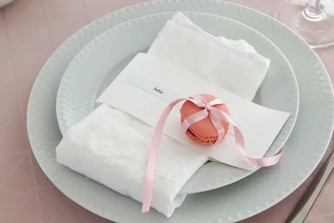 To hvite tallerker, kniv og gaffel, rosa duk og hvit serviett med rosa makron knyttet med silkebånd og en navnelapp hvor de står Julie