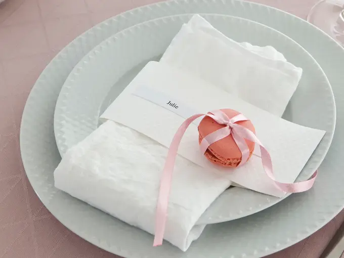 To hvite tallerker, kniv og gaffel, rosa duk og hvit serviett med rosa makron knyttet med silkebånd og en navnelapp hvor de står Julie
