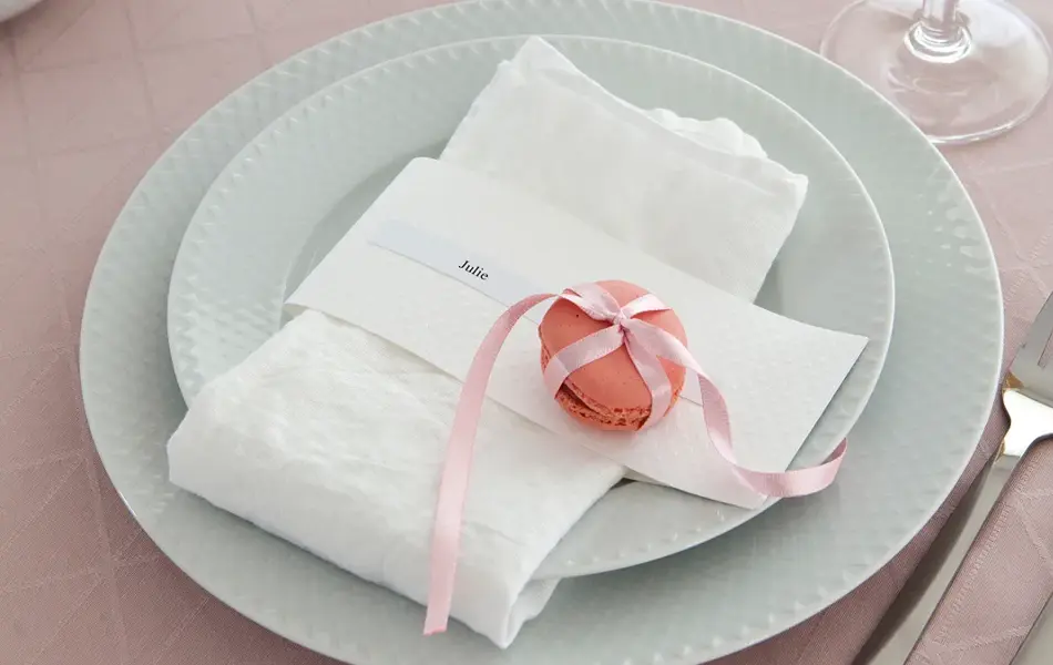 To hvite tallerker, kniv og gaffel, rosa duk og hvit serviett med rosa makron knyttet med silkebånd og en navnelapp hvor de står Julie
