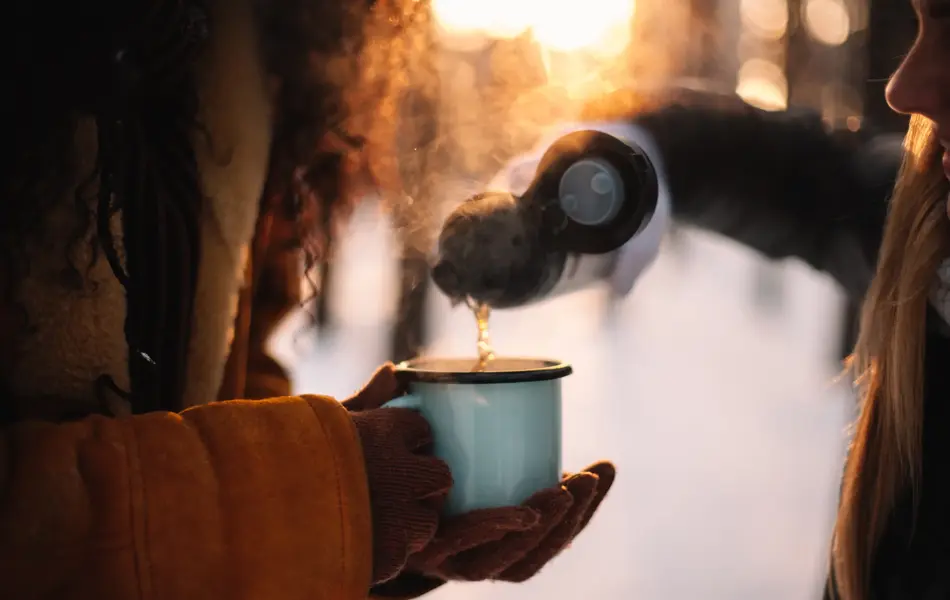 Woman pouring tea for girlfriend in cup standing at park during winter