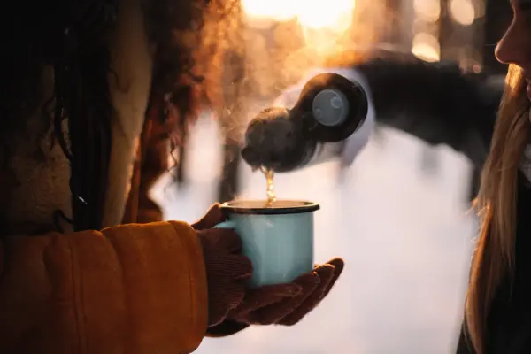 Woman pouring tea for girlfriend in cup standing at park during winter