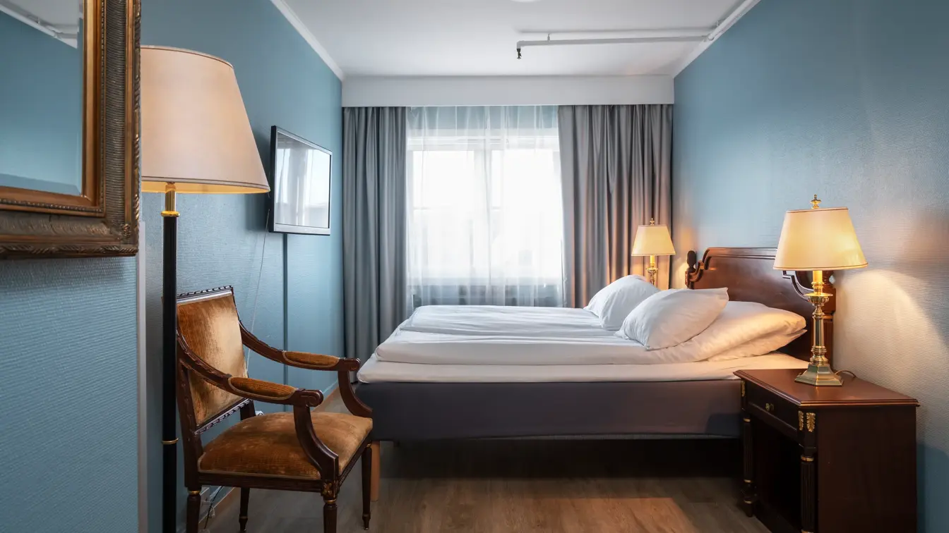 Dobbeltseng, nattbord, vindu, grå gardiner, stol, parkettgulv, turkise vegger. 