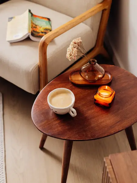 Lite brunt bord med tent stearinlys, blomst i krukke og kaffekopp med hvit stol med en oppslått bok i bakgrunnen 