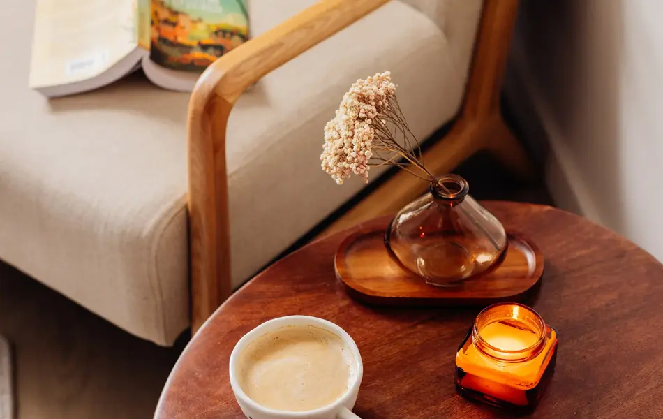 Lite brunt bord med tent stearinlys, blomst i krukke og kaffekopp med hvit stol med en oppslått bok i bakgrunnen 