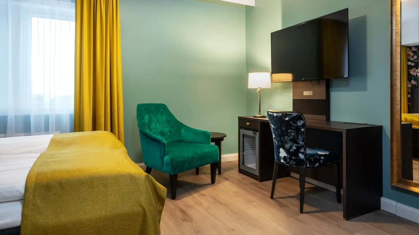 Turkise vegger, mørkt skrivebord med veggehengt TV, og mønstrete stol. Grønn lenestol. Vindu med gul gardin. Dobbeltseng med gult sengeteppe. I dobbeltrom på Thon Hotel Høyers i Skien.