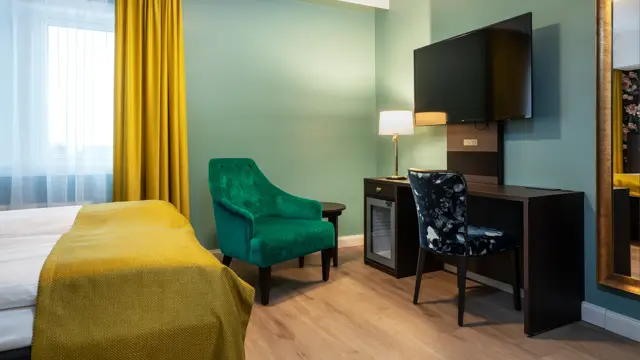Turkise vegger, mørkt skrivebord med veggehengt TV, og mønstrete stol. Grønn lenestol. Vindu med gul gardin. Dobbeltseng med gult sengeteppe. I dobbeltrom på Thon Hotel Høyers i Skien.