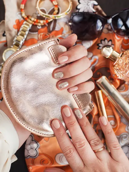 Et nærbilde av to damehender med metallisk neglelakk som holder en liten metallisk veske.