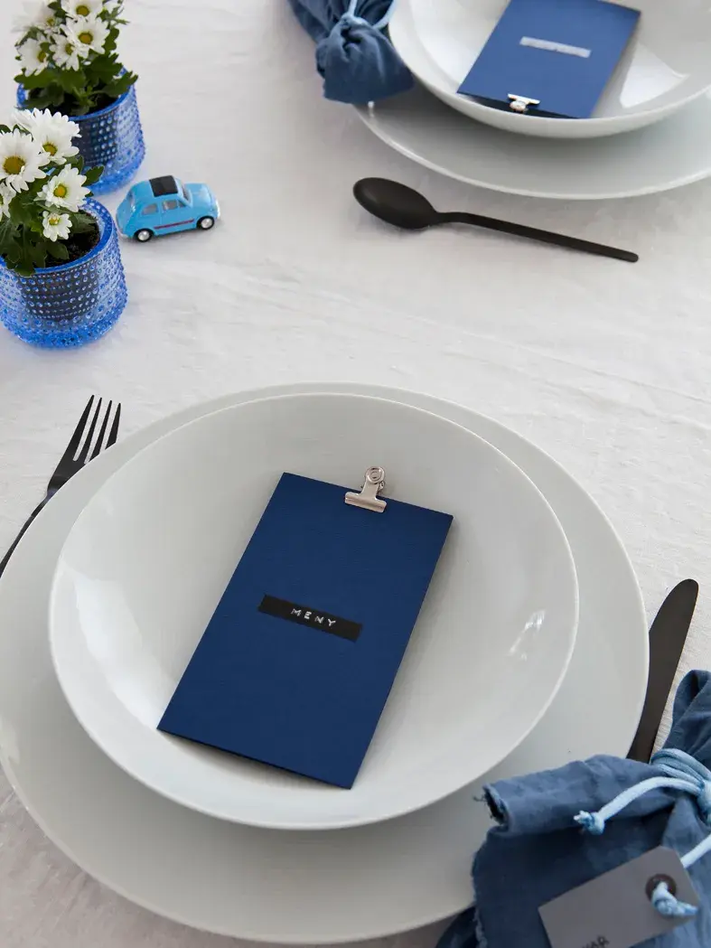 Steg 5 - bord dekket med mørkeblå tøyservietter og blått menykort, hvite blomster og en liten lekebil 