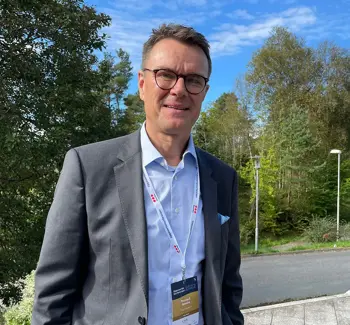 Halvtotal av dresskledd Thomas Rønning som smiler mot kamera, med vei og skogområde i bakgrunnen.