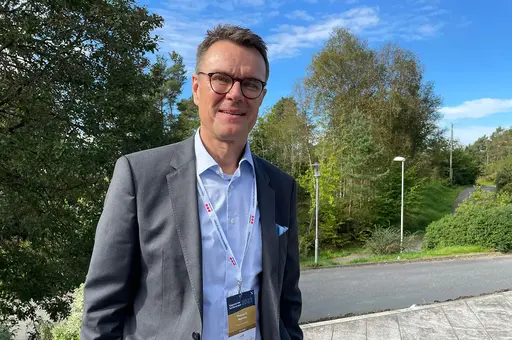 Halvtotal av dresskledd Thomas Rønning som smiler mot kamera, med vei og skogområde i bakgrunnen.