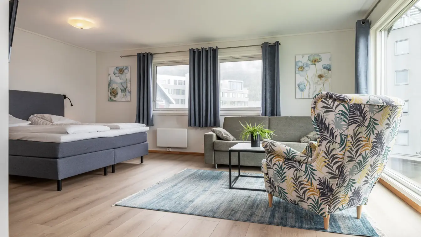 Superior rom med dobbeltseng, store vinduer, sofa, stor lenestol, parkett med et stort firkantet teppe under sittegruppe.