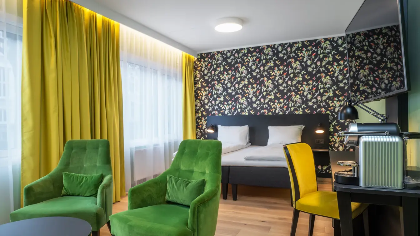 Dobbeltseng, mønstrete tapetvegg, lysegrønne vegger, TV, skrivebord, gul stol, kaffemaskin,  to grønne stoler, parkettgulv,vindu fra gulv til tak, gule gardiner, svart salongbord. 
