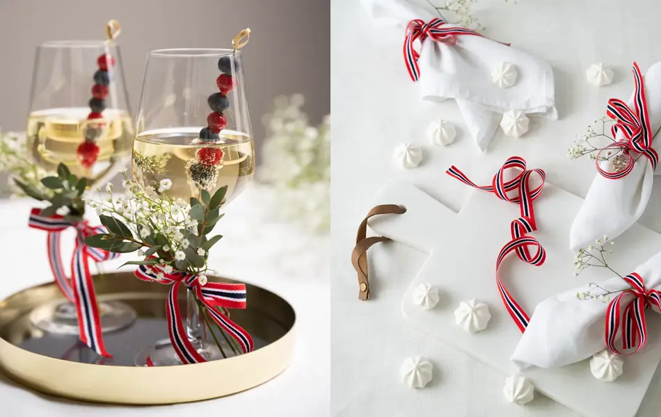 To vinglass pyntet med 17.mai-bånd og blomster. Hvit duk med hvite servietter med 17.mai-bånd og hvite marengs på bordet.
