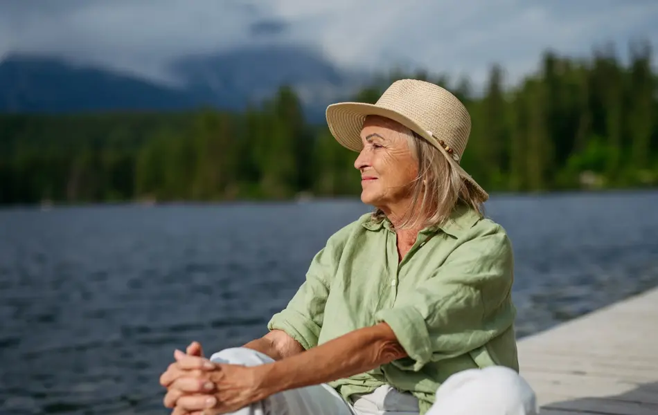 Eldre dame med pastellgrønn skjorte og stråhatt sitter på bryggekant og ser utover vannet