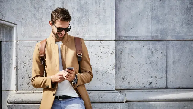 Mann med solbriller og lysebrun slim jakke ser på mobilen foran murvegg 