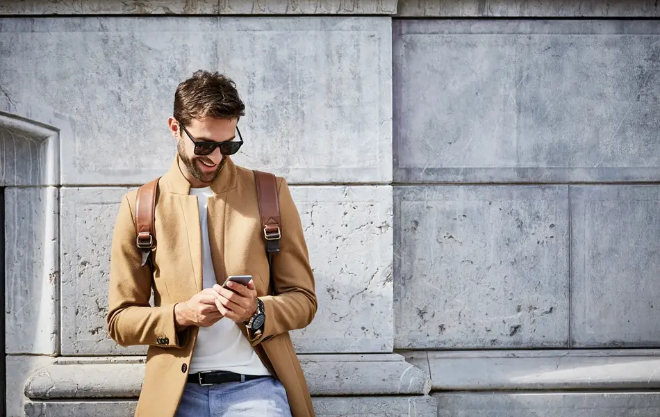 Mann med solbriller og lysebrun slim jakke ser på mobilen foran murvegg 
