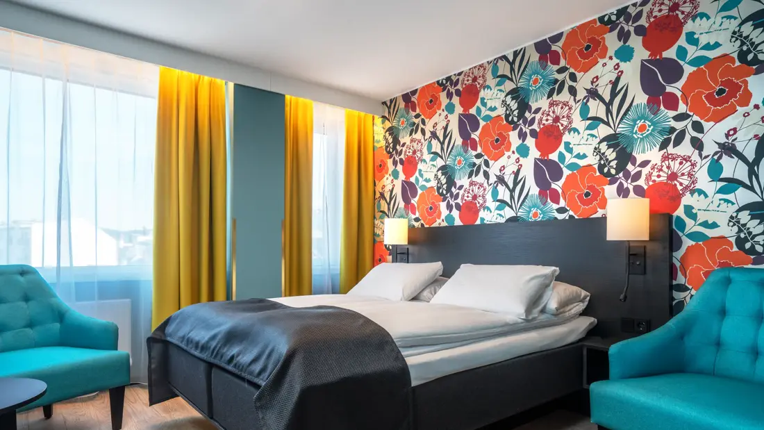 Blomstertapetvegg, dobbeltseng, turkis sofa, turkis stol, salongbord, parkettgulv, vindu, gule gardiner. 