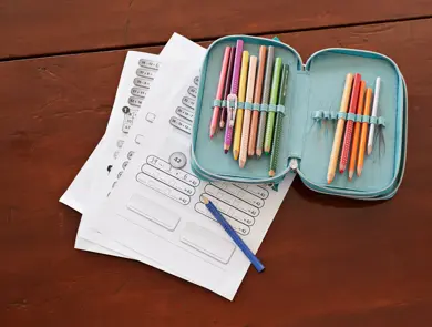 grønn pennal med fargeblyanter som ligger oppå skoleoppgaver