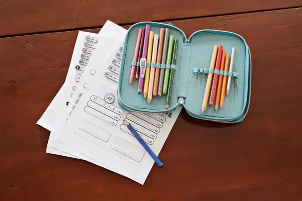 grønn pennal med fargeblyanter som ligger oppå skoleoppgaver