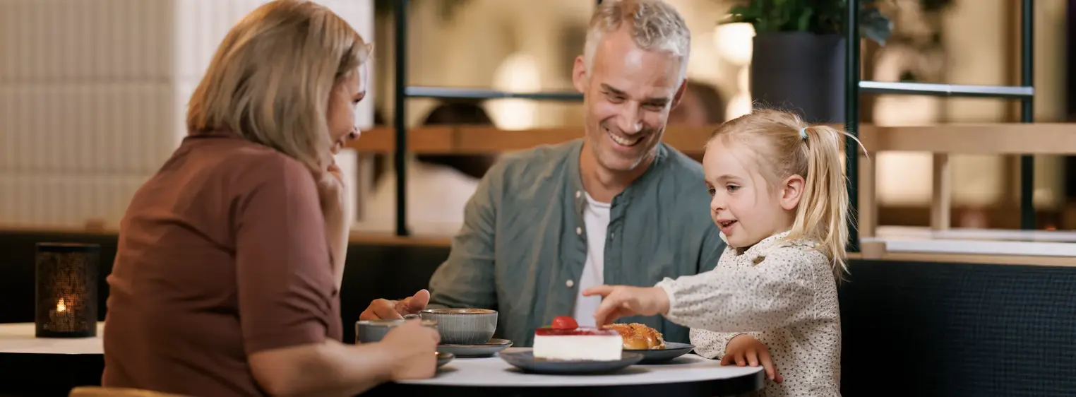 En mann, en kvinne og et barn sitter ved et bord på en kafé, mannen og kvinnen smiler til barnet