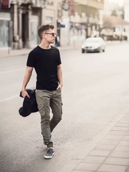 Mann gående over gata iført grønn cargobukse, svart t-skjorte og solbriller.