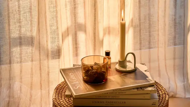 Lite bord med magasiner og stearinlys på, foran et vindu med florlette gardiner.