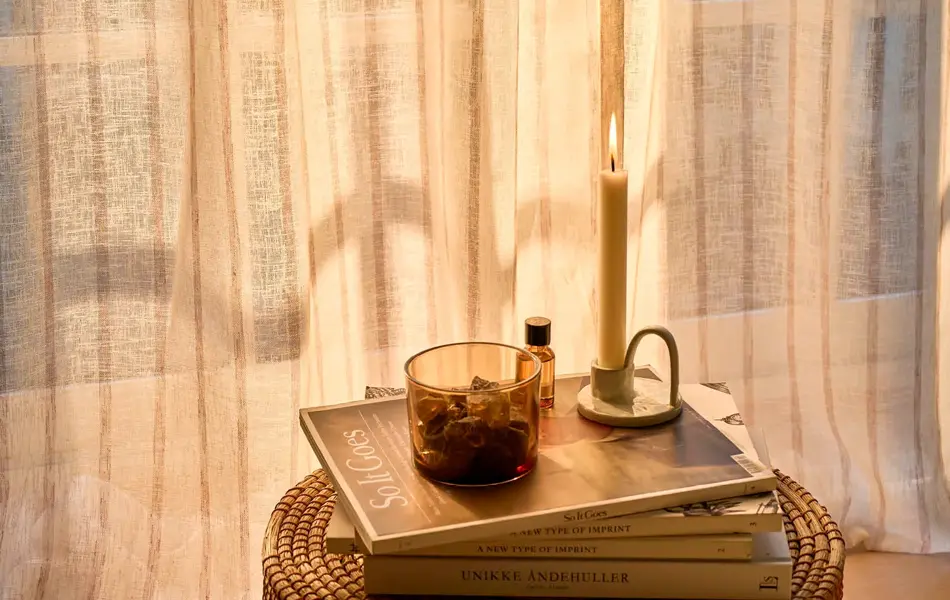 Lite bord med magasiner og stearinlys på, foran et vindu med florlette gardiner.