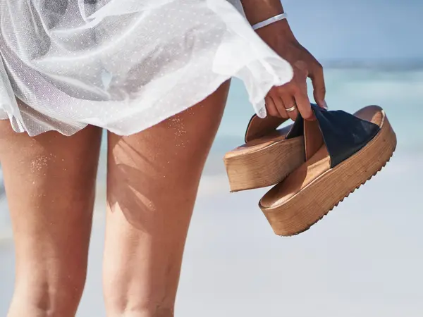 Kvinne står vendt mot havet med et par sandaler med store såler i hånda