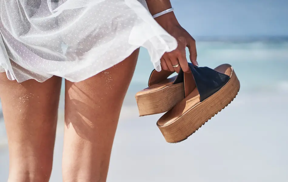 Kvinne står vendt mot havet med et par sandaler med store såler i hånda