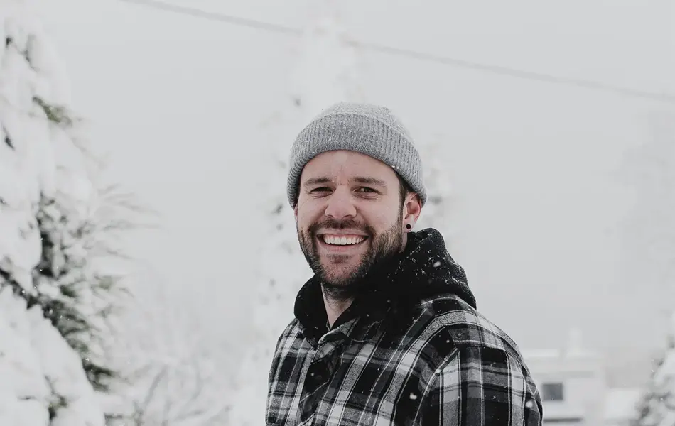 Mann med svart flanellskjorte og grå lue smiler til kamera med snødekt landskap i bakgrunn 