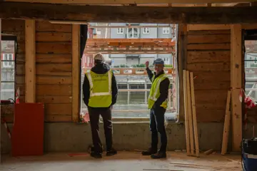Trond Are og Ståle ved vindu mot Nidelva i 1. etasje i Kjøpmannsgata 15 i Trondheim.  Begge i hjelm og gul vest, oppussing og diverse materialer rundt.