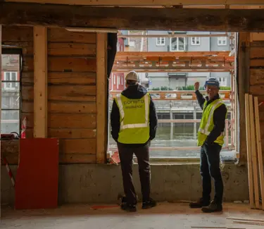 Trond Are og Ståle ved vindu mot Nidelva i 1. etasje i Kjøpmannsgata 15 i Trondheim.  Begge i hjelm og gul vest, oppussing og diverse materialer rundt.