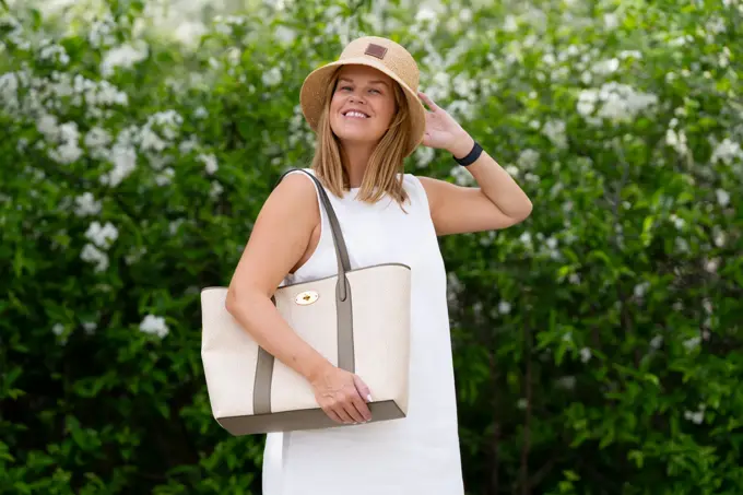 Damemodell står ute med hvit kjole lys beige strandveske og stråhatt