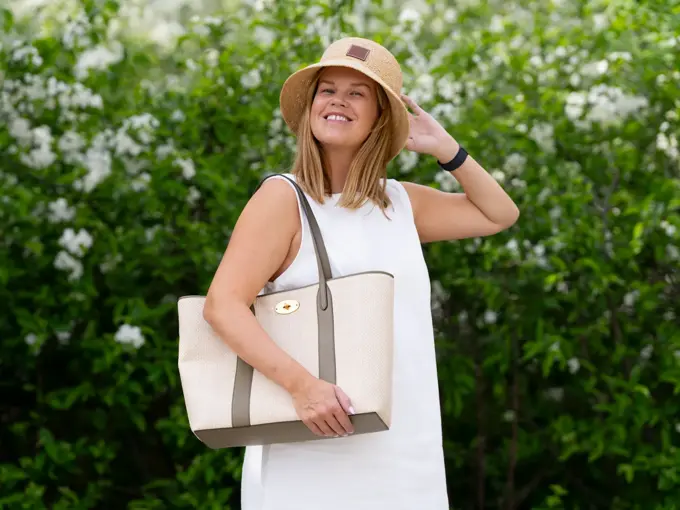 Damemodell står ute med hvit kjole lys beige strandveske og stråhatt