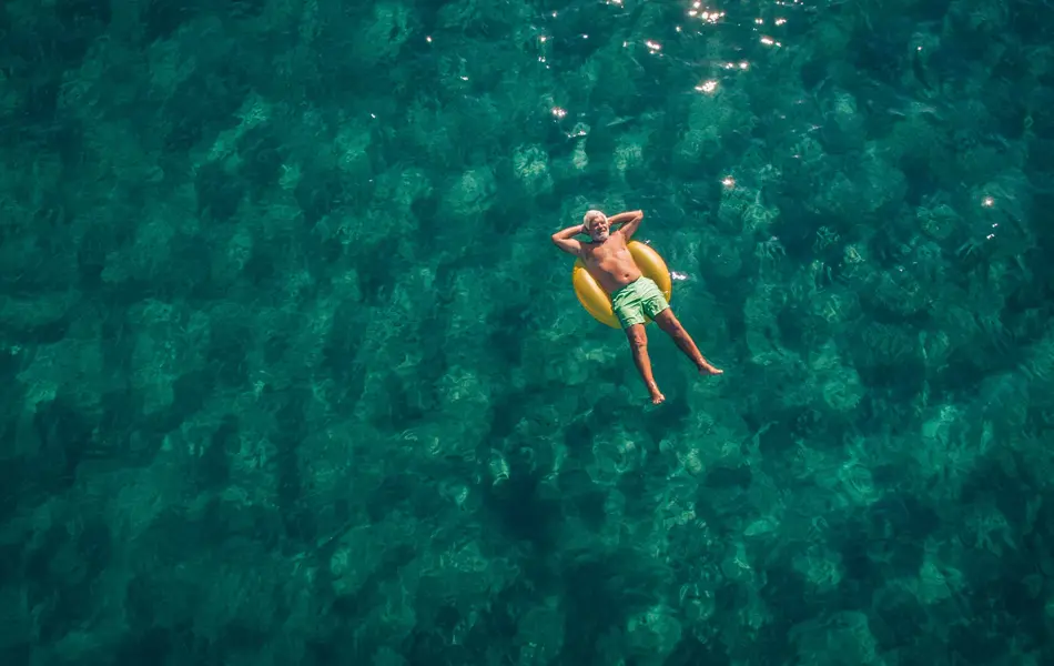 Bilde tatt ovenfra av en eldre mann iført en grønn badebukse. Mannen ligger på en gul badering i sjøen, med armene under hodet.  