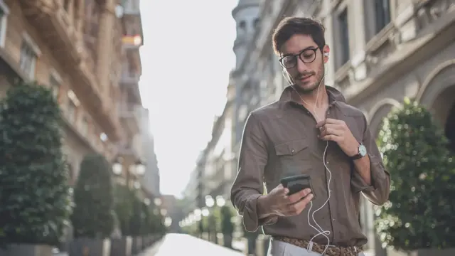 Mann i by iført brun skjorte, beige chinos og flettet belte. Briller, ørepropper med kabel, mobil.