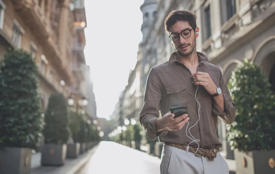 Mann i by iført brun skjorte, beige chinos og flettet belte. Briller, ørepropper med kabel, mobil.