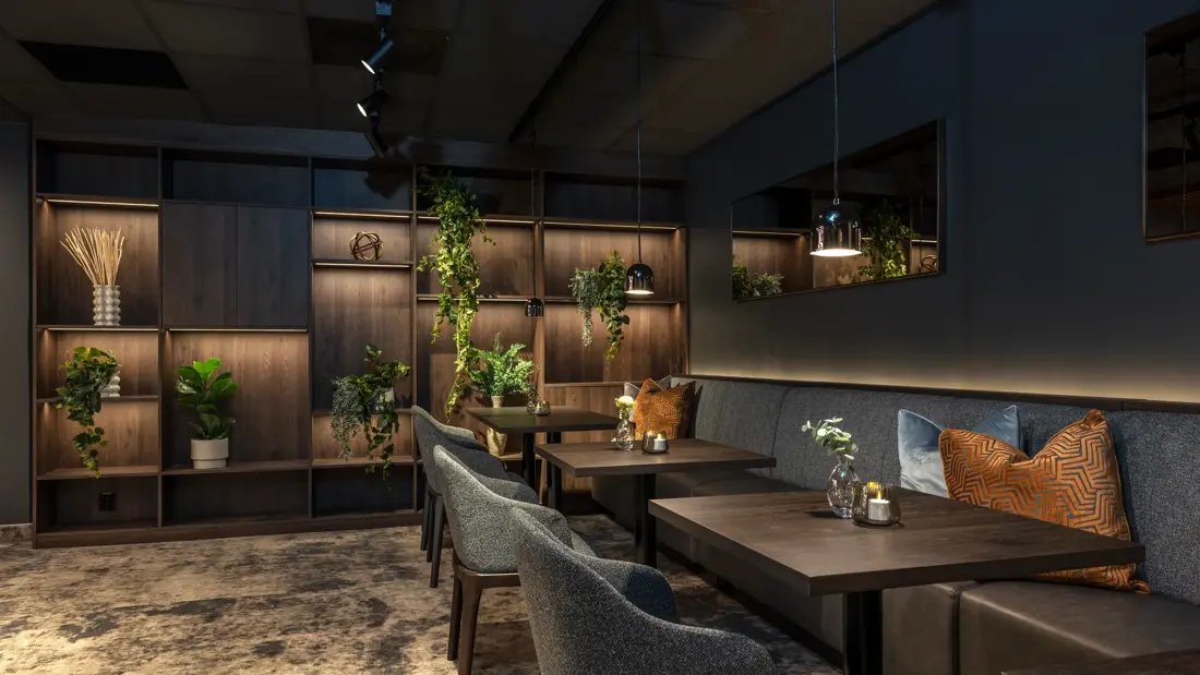 Deler av frokostsal med bord, stoler, brun bokhylle bakerst med diverse planter og pynt. Teppegulv og dempet belysning.