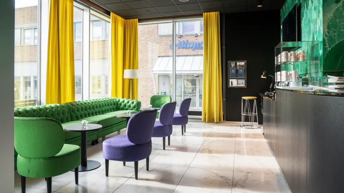 Lyst gulv, svart tak, grønn sofa, tre lilla stoler, en grønn stol, vinduer fra tak til gulv, gule gardiner, kaffestasjon med kaffekopper. 