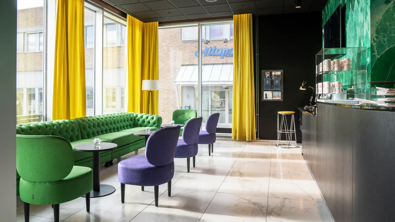 Lyst gulv, svart tak, grønn sofa, tre lilla stoler, en grønn stol, vinduer fra tak til gulv, gule gardiner, kaffestasjon med kaffekopper. 