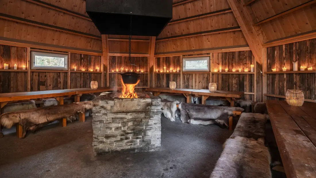 Lavvo med dempet belysning, trebord, skinn på benkene og bål i midten.