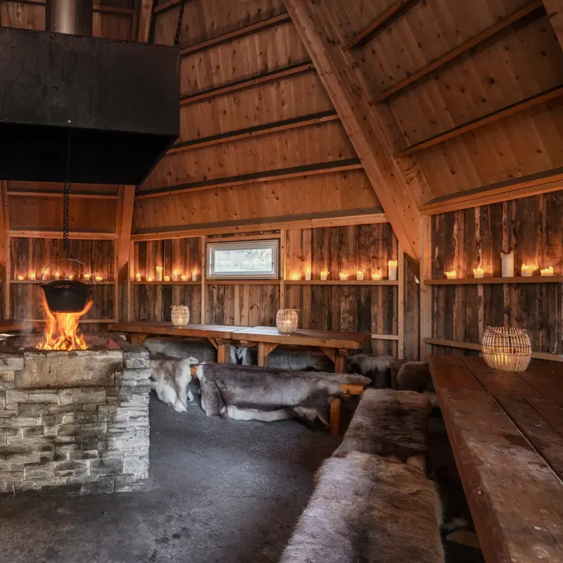 Lavvo med dempet belysning, trebord, skinn på benkene og bål i midten.