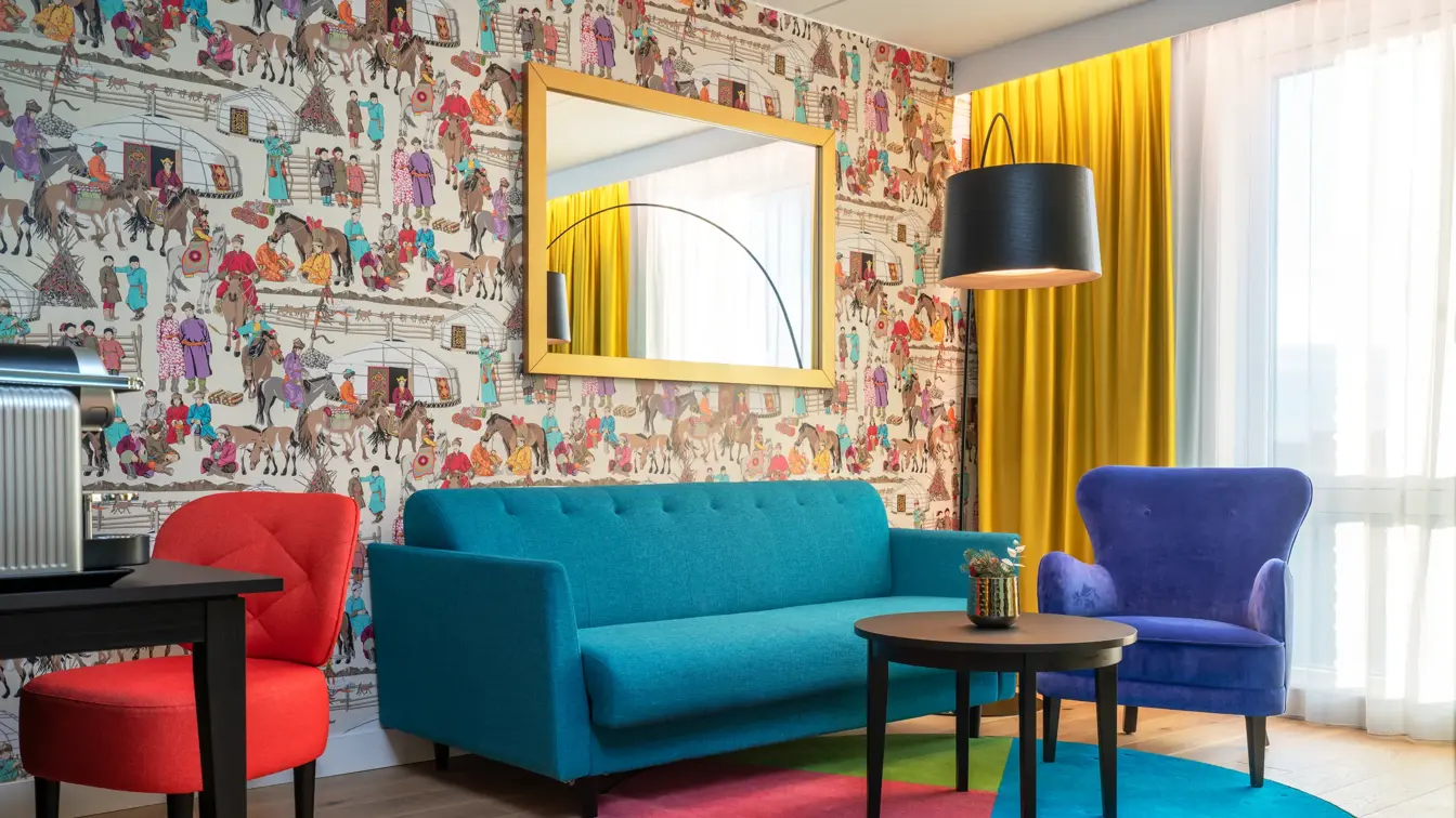 Dekorativ tapet, speil med gullramme, sittegruppe med turkis sofa, blå stol og bord på fargerikt teppe i suite  på Thon Hotel Storo
