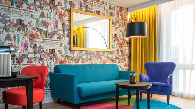 Dekorativ tapet, speil med gullramme, sittegruppe med turkis sofa, blå stol og bord på fargerikt teppe i suite  på Thon Hotel Storo