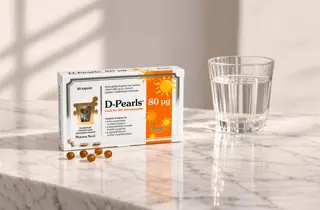 En boks med D-Pearls fra Pharma Nord står ved siden av et glass med vann.